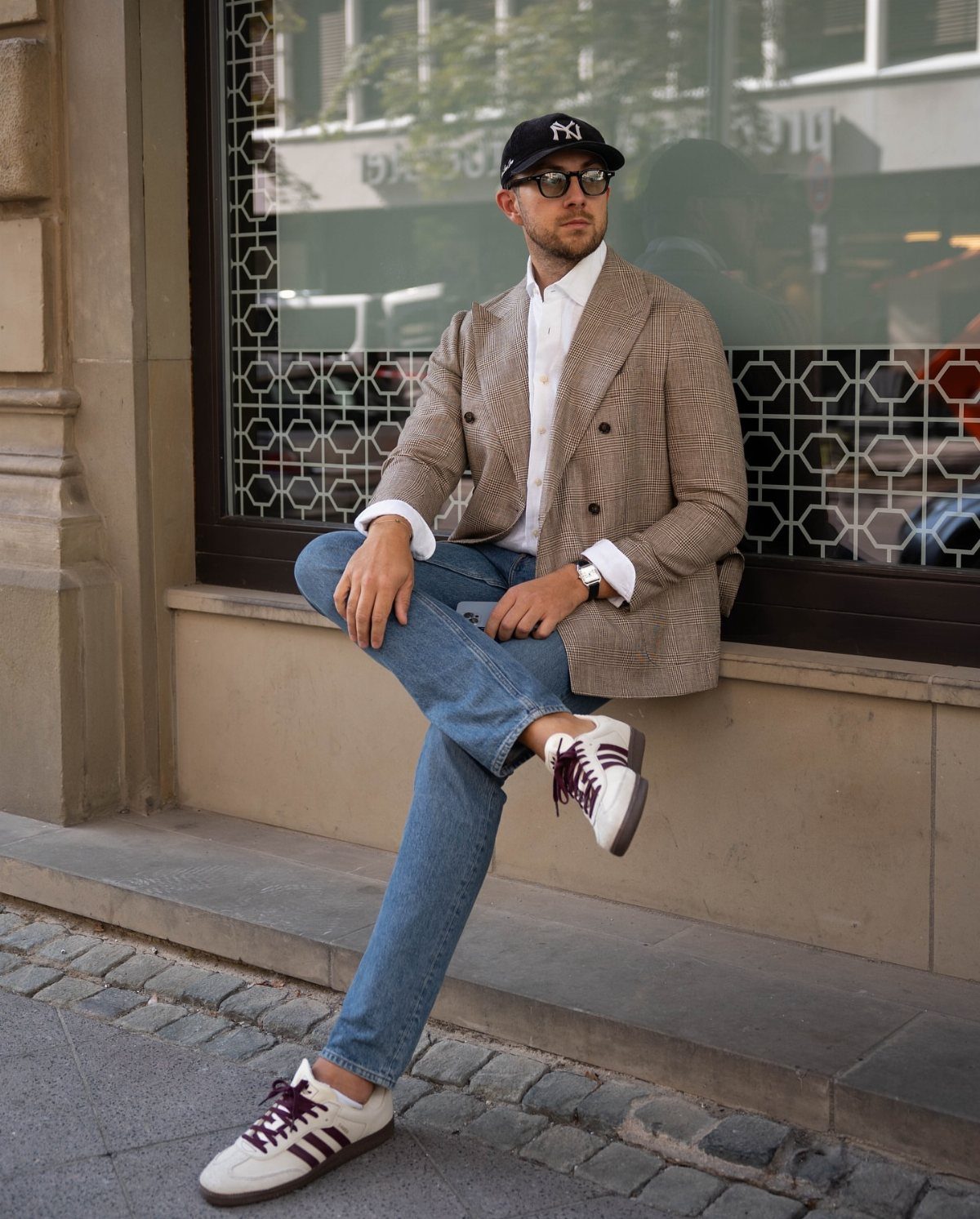 A man wearing a checkered blazer, blue jeans, and white sneakers sitting on a stone ledge.