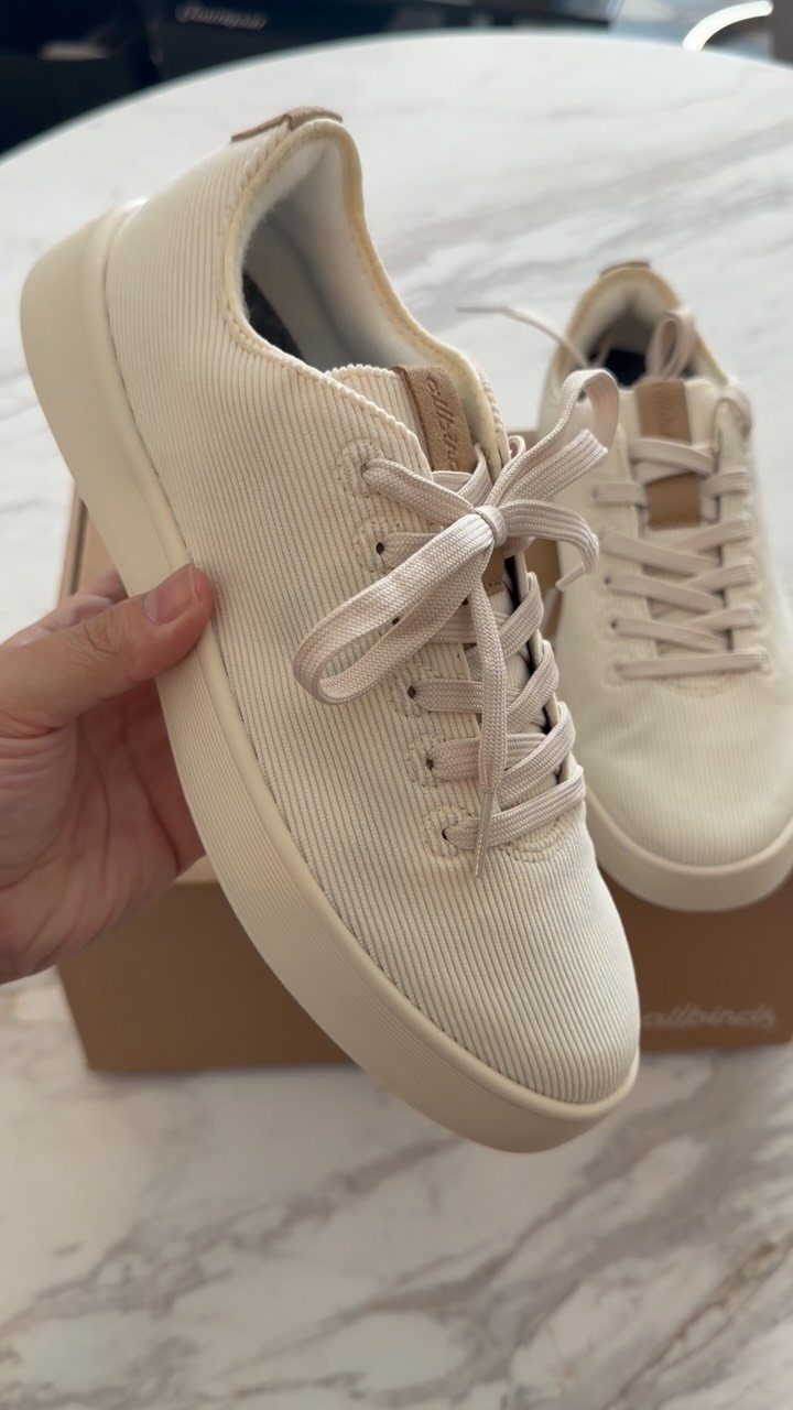 A hand holding a cream-colored corduroy sneaker over a marble tabletop.