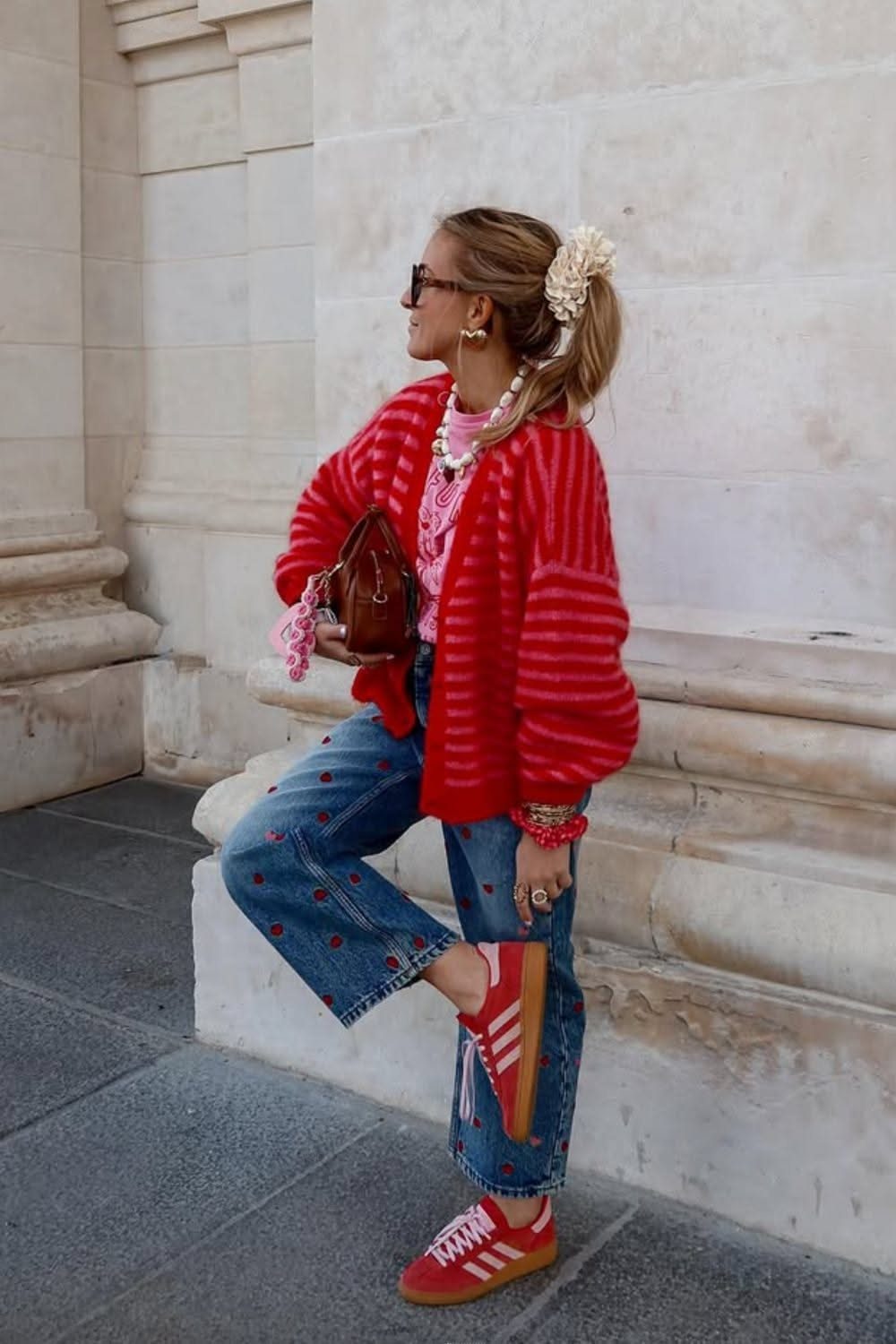 Woman wearing a red oversized cardigan, blue embroidered jeans, and red sneakers standing by a stone wall.