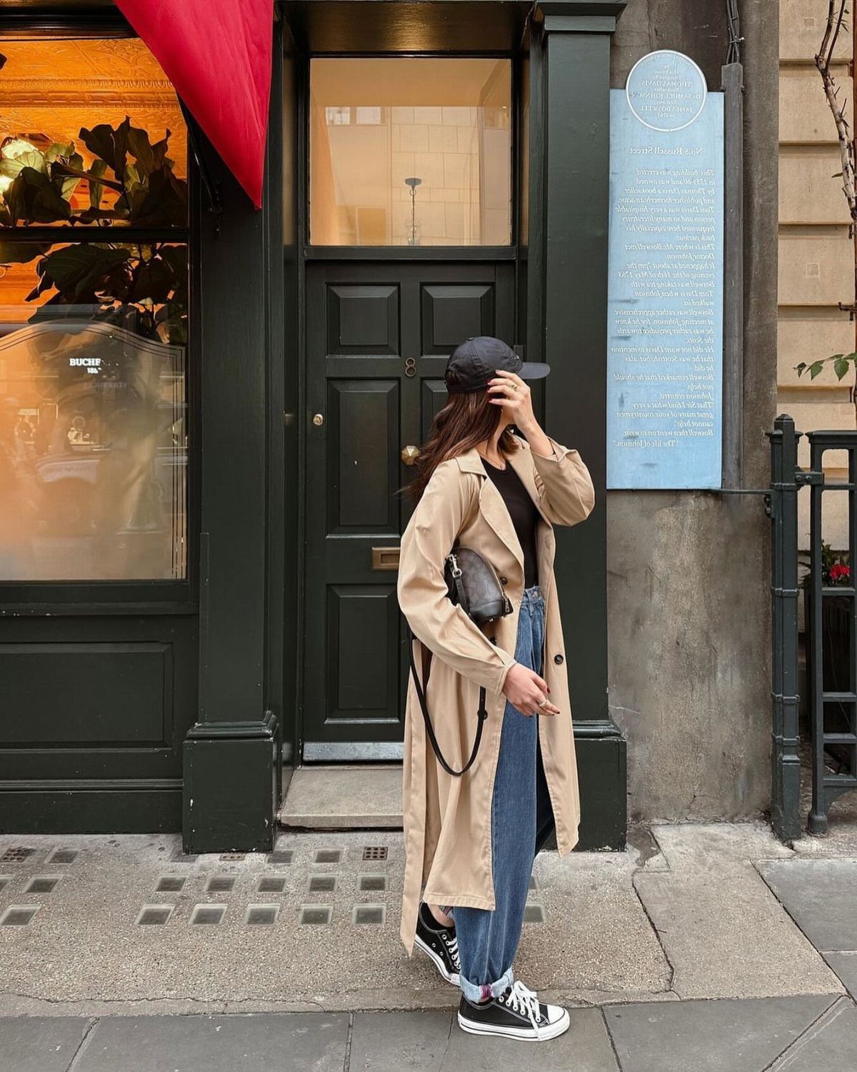 A woman wearing a tan trench coat, blue jeans, and black sneakers standing on a city street.