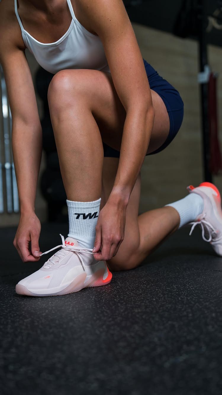 A person kneeling on a gym floor tying the laces of a light pink athletic sneaker.