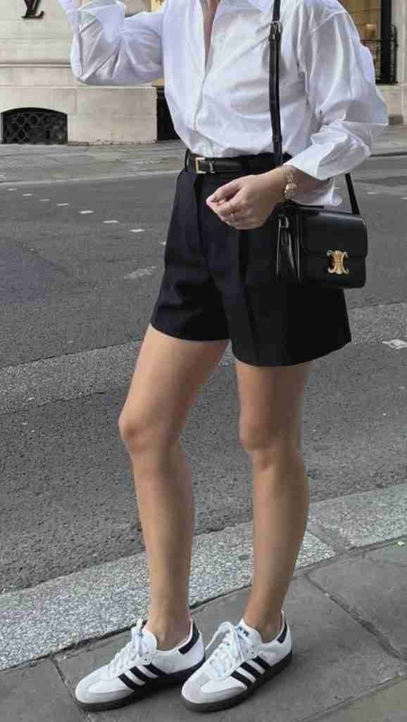 Person wearing white button-down shirt, black shorts, and white Adidas sneakers standing on a street.