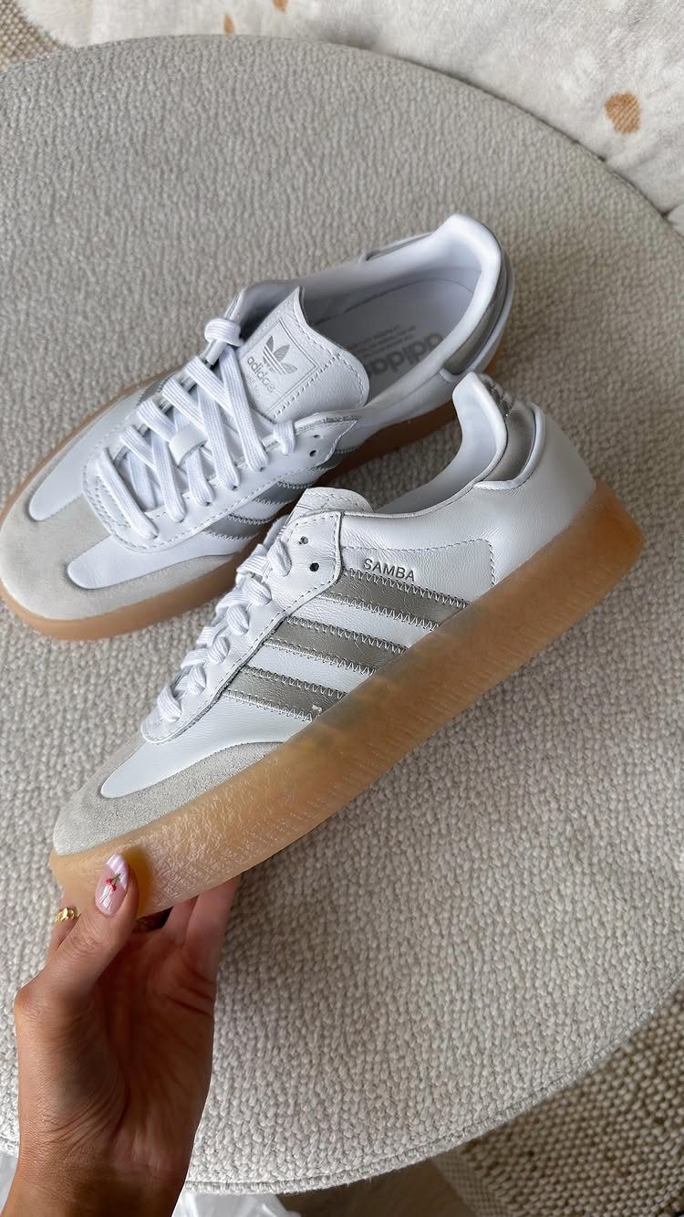 A hand holds a pair of white Adidas Samba sneakers with silver stripes and gum soles.