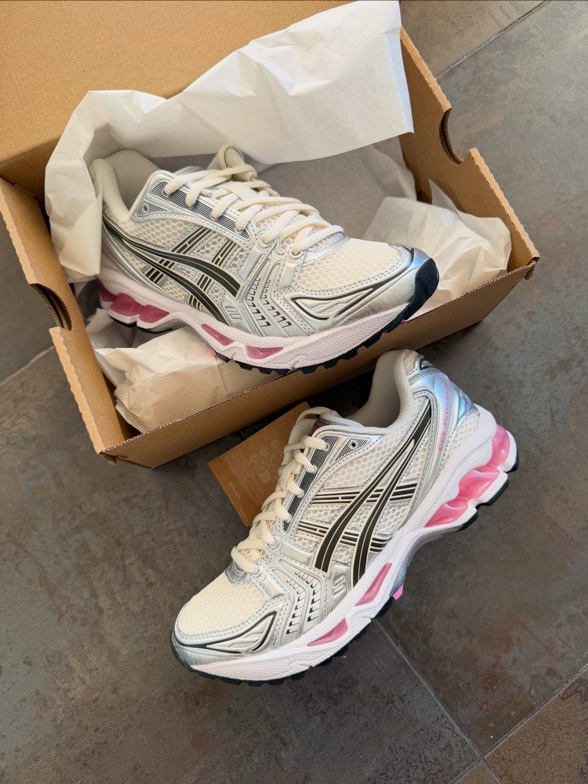 White and silver Asics Gel-Kayano 14 sneakers with pink accents sitting on a grey floor.