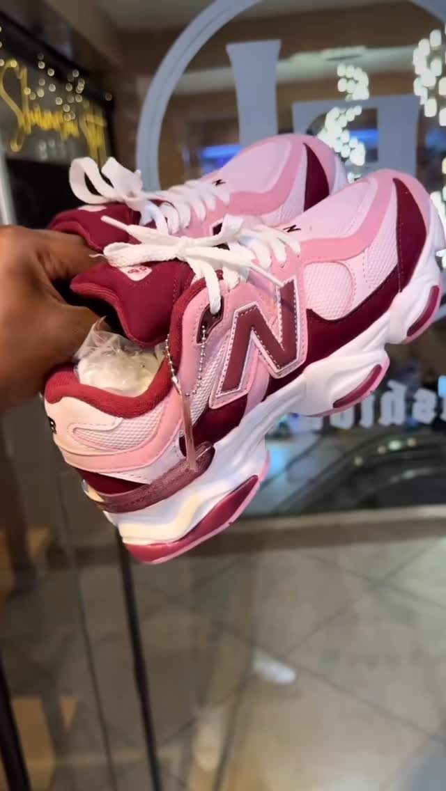 A hand holding pink and burgundy New Balance chunky sneakers against a glass background.