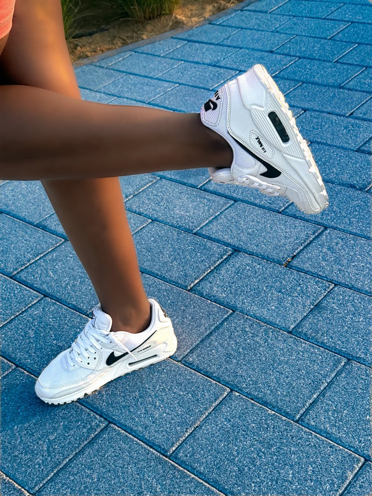 White Nike Air Max sneakers worn by a person walking on blue paving stones.