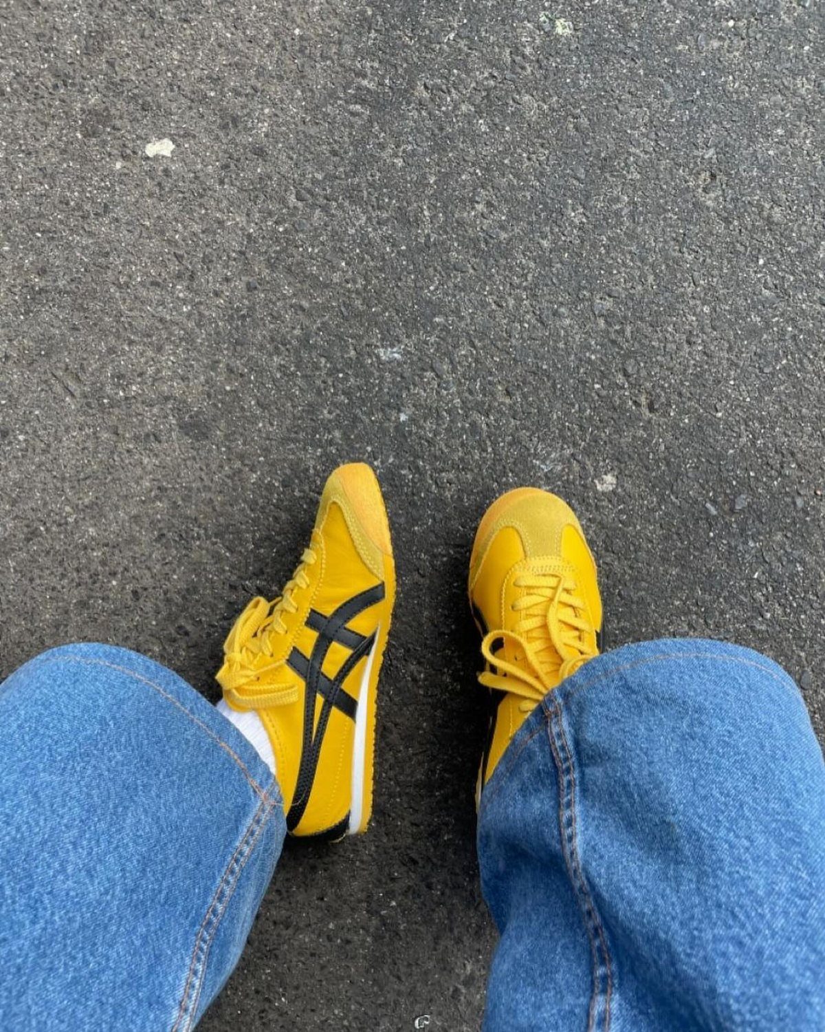 Yellow suede sneakers with black stripes worn with blue jeans on asphalt.