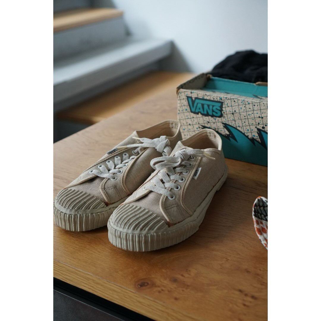 Beige canvas plimsoll sneakers with thick ribbed soles resting on a wooden table near a shoebox.