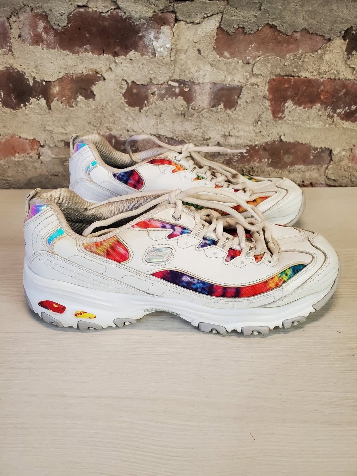 White Skechers athletic shoes with vibrant tie-dye patterns on the sides sitting on a wooden surface.