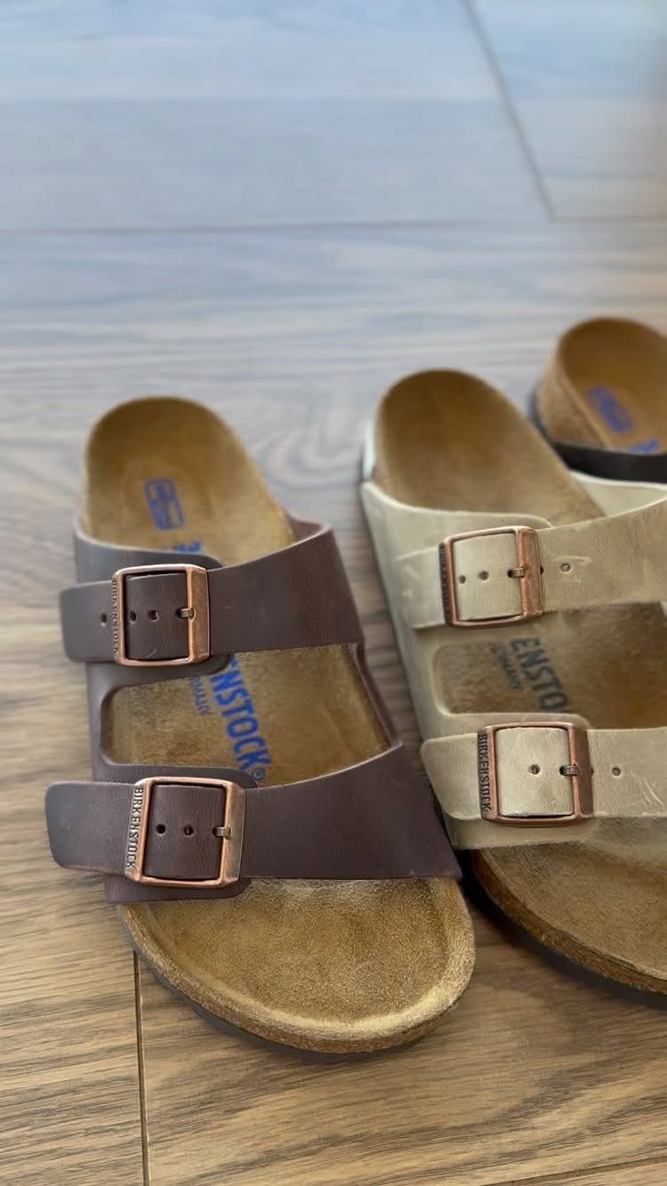 Two pairs of two-strap leather sandals with cork footbeds resting on a wooden floor.