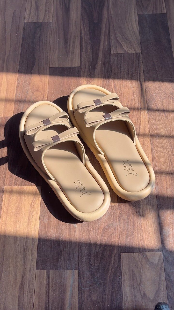 Beige platform slide sandals with bow-shaped straps on a wooden floor.