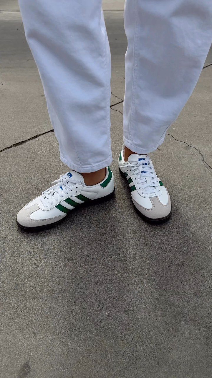 Person wearing white trousers and green striped Adidas sneakers on a concrete ground.