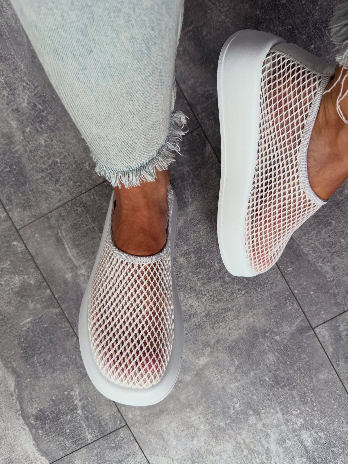 Person wearing white mesh knit slip-on sneakers and light blue frayed denim jeans on grey tiles.