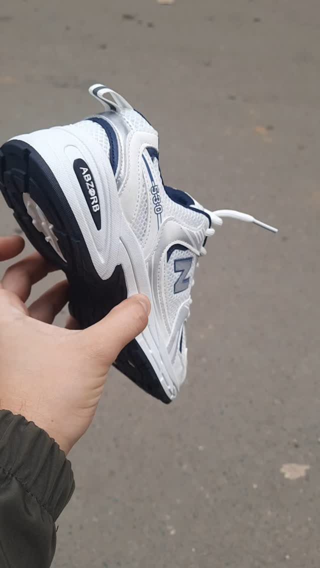 A hand holding a white and navy New Balance 530 sneaker against a grey pavement background.