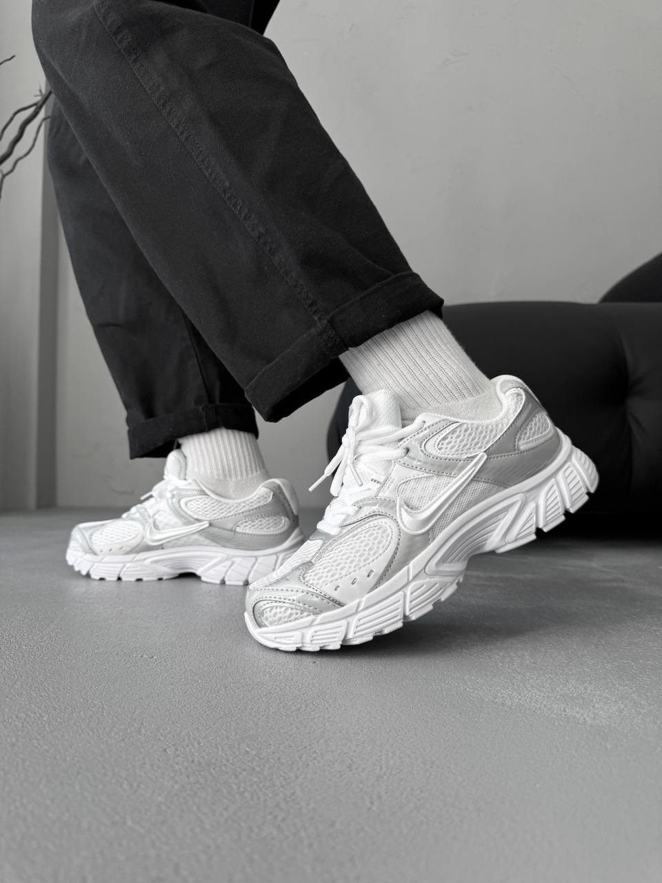 A person wearing white Nike sneakers with mesh details and black trousers on a grey floor.