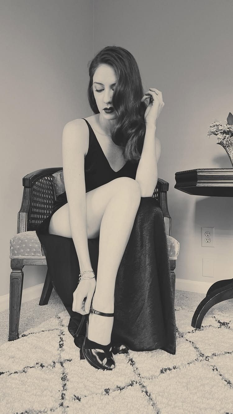 A woman sitting in a chair while putting on a black peep-toe ankle strap high heel.