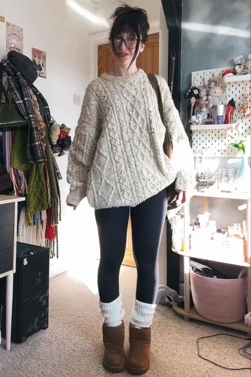 A person wearing a cream cable-knit oversized sweater, dark leggings, white leg warmers, and tan boots.