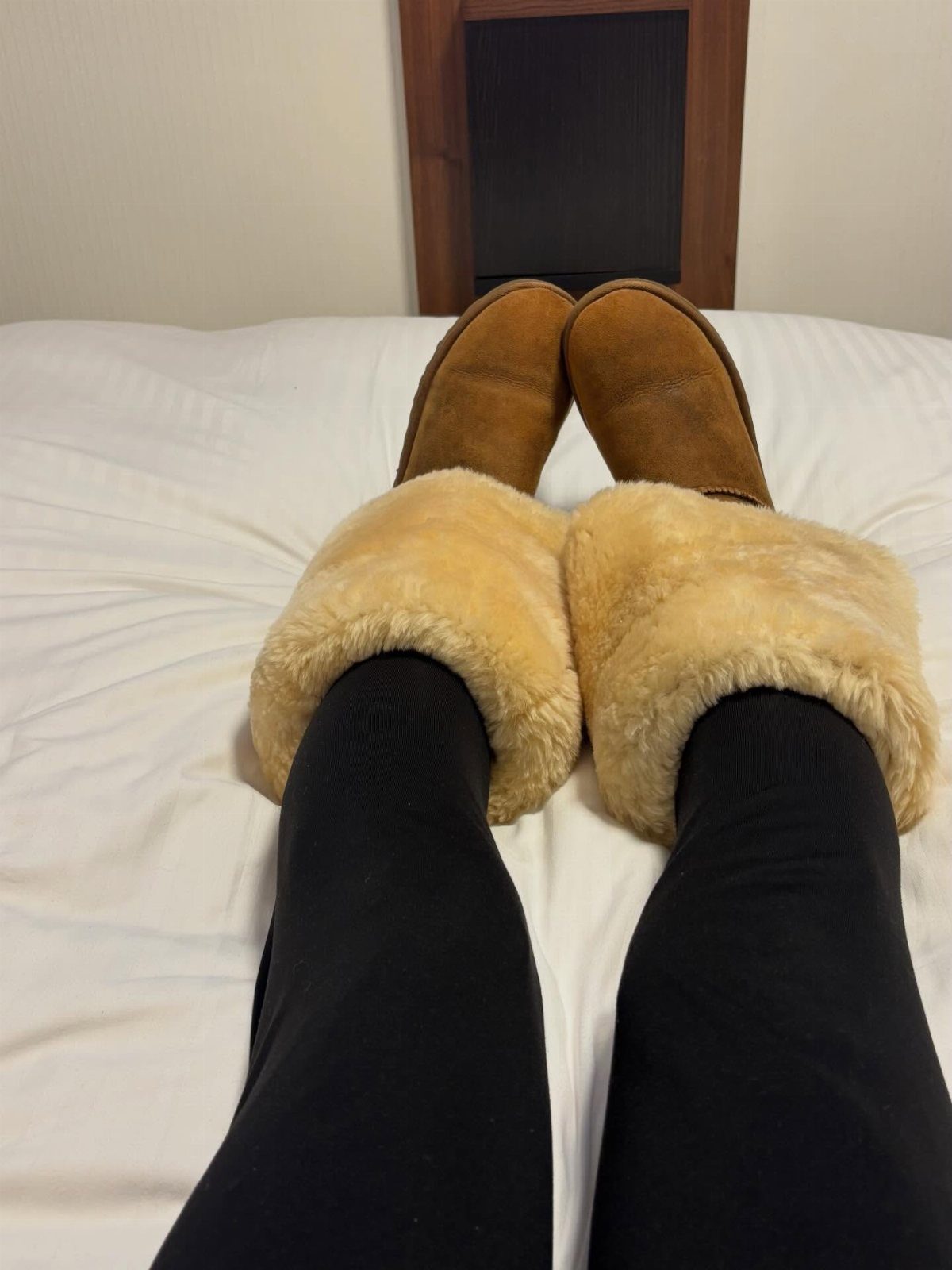 Tan suede boots with fluffy cream leg warmers and black leggings on a white bed.