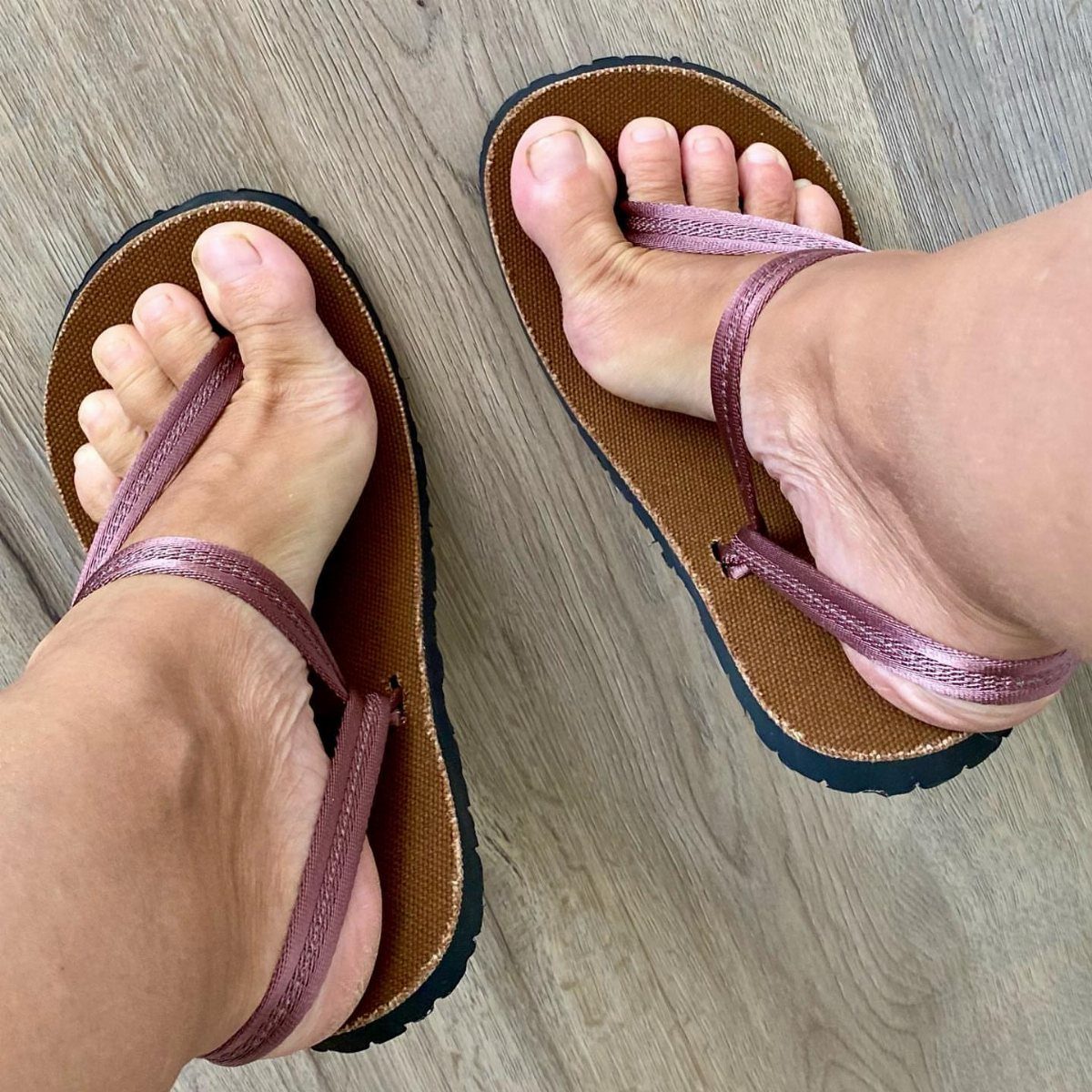 A person wearing pink thong sandals with brown soles while standing on a wooden floor.