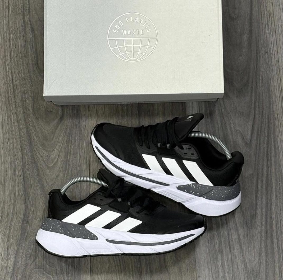 Black Adidas sneakers with white stripes and thick white soles on a wooden floor.
