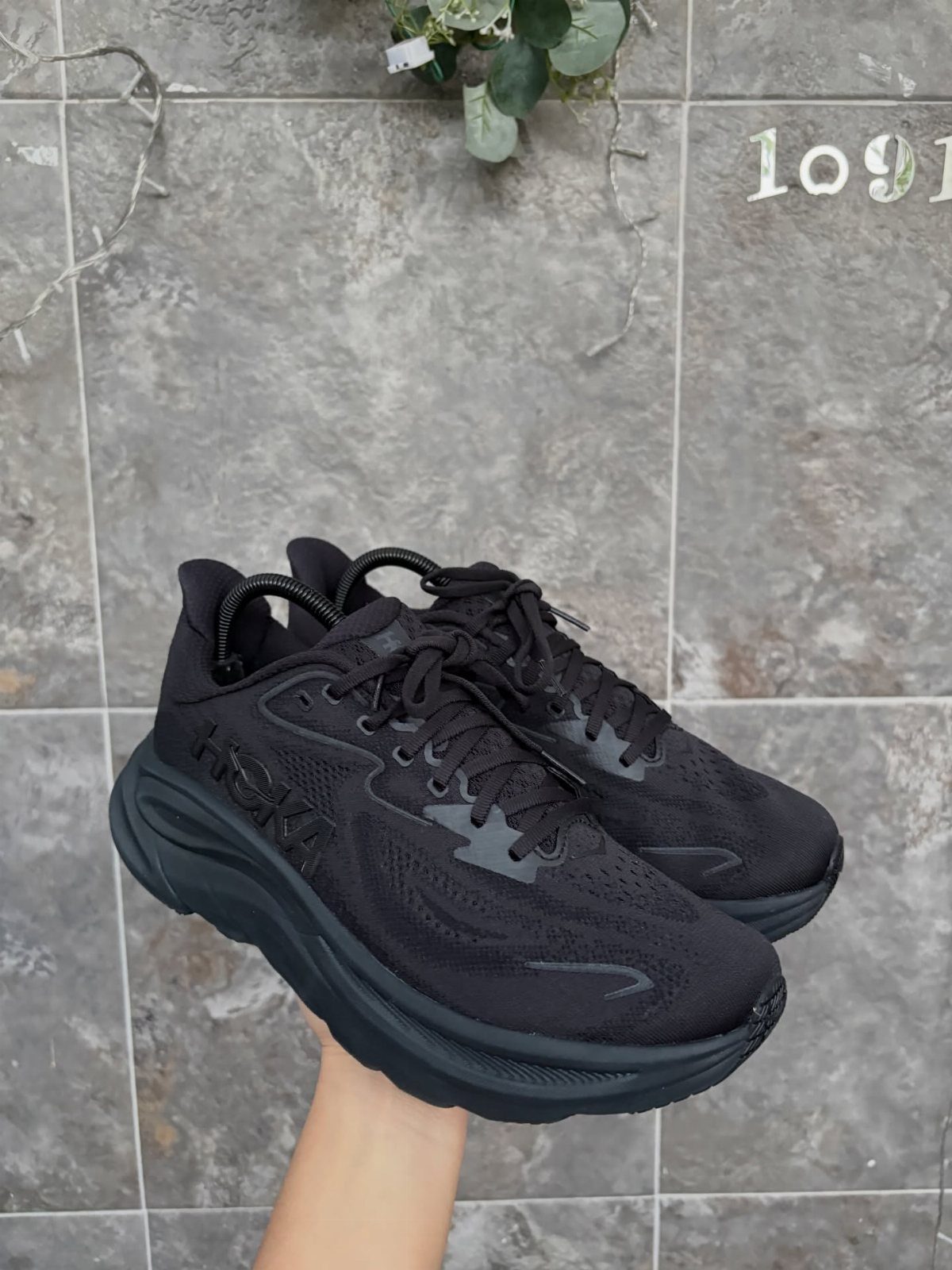 A hand holding up a pair of black athletic walking shoes against a grey tiled wall.