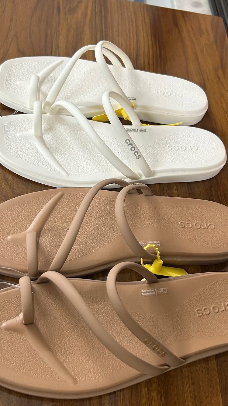 Two pairs of Crocs toe loop sandals in white and tan on wood.