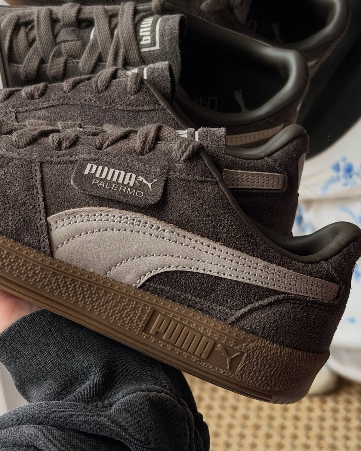 Dark grey suede Puma Palermo sneakers with light grey stripes and a brown gum sole being held.