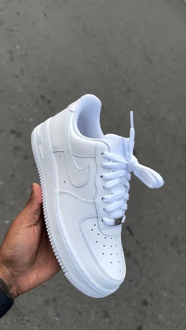 A hand holding a white Nike Air Force 1 sneaker against a grey pavement background.