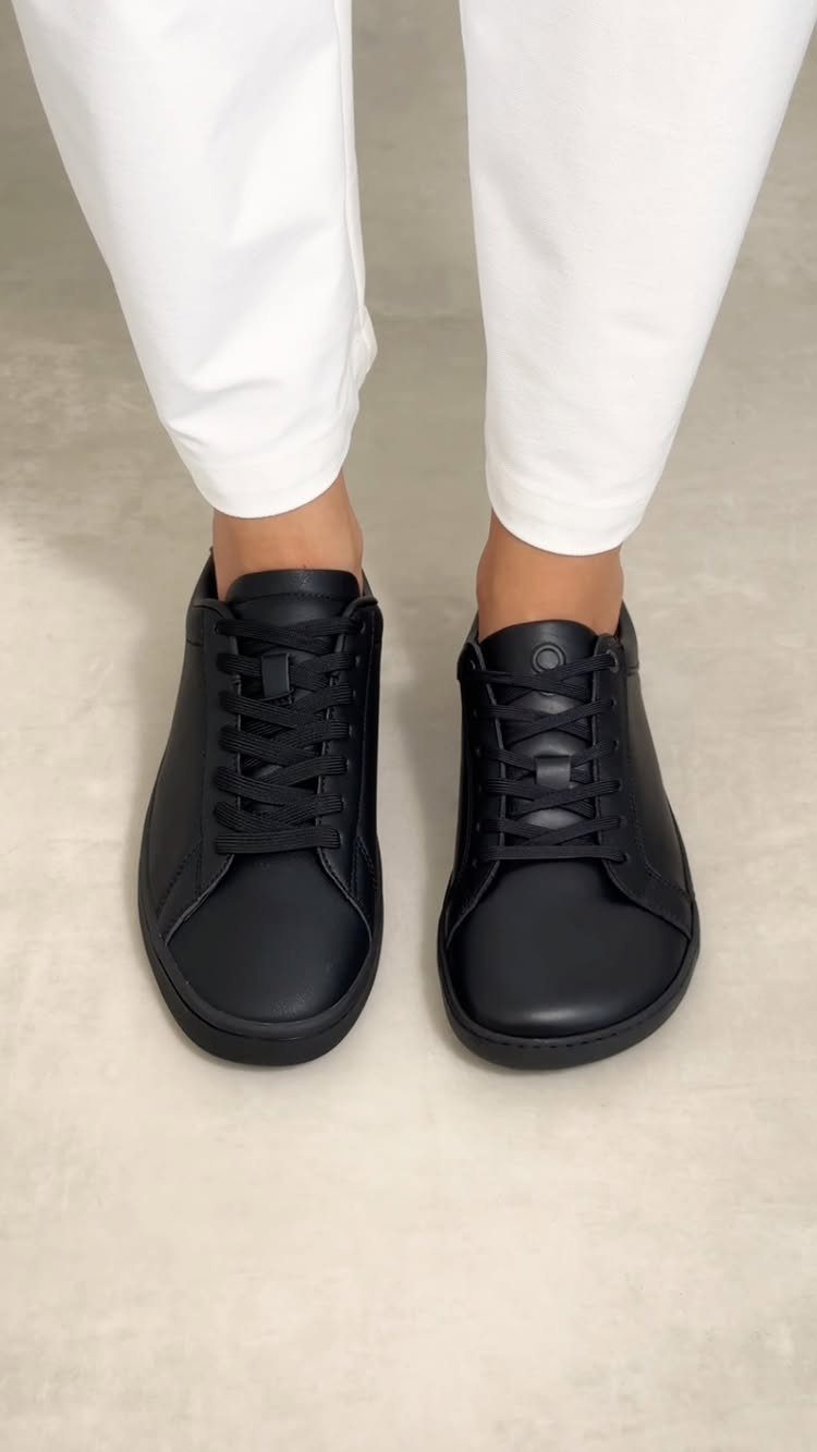 Black leather low-top sneakers worn by a person with white trousers on a grey floor.